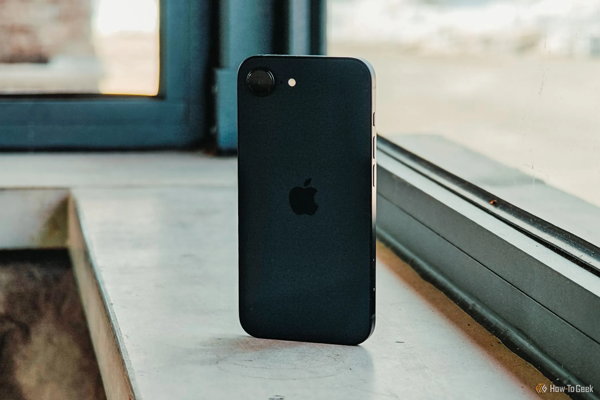 Mặt lưng iPhone 16e màu trắng nhìn từ góc nghiêng, đặt trước khung cửa sổ đón ánh sáng tự nhiên