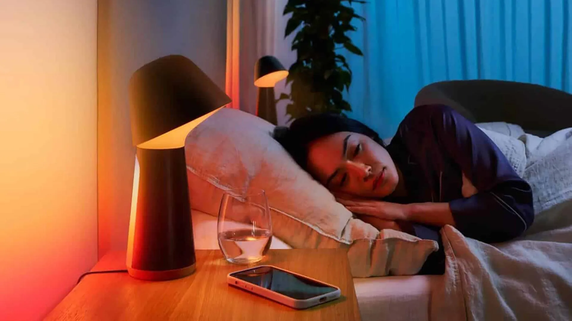 Một người phụ nữ đang ngủ say trên giường bên cạnh chiếc đèn thông minh và điện thoại thông minh, minh họa việc sử dụng điện thoại để tạo tiếng ồn trắng hỗ trợ giấc ngủ.