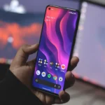 Người dùng cầm điện thoại Android trên bàn làm việc có nhiều màn hình máy tính, minh họa trải nghiệm điều hướng trên thiết bị di động