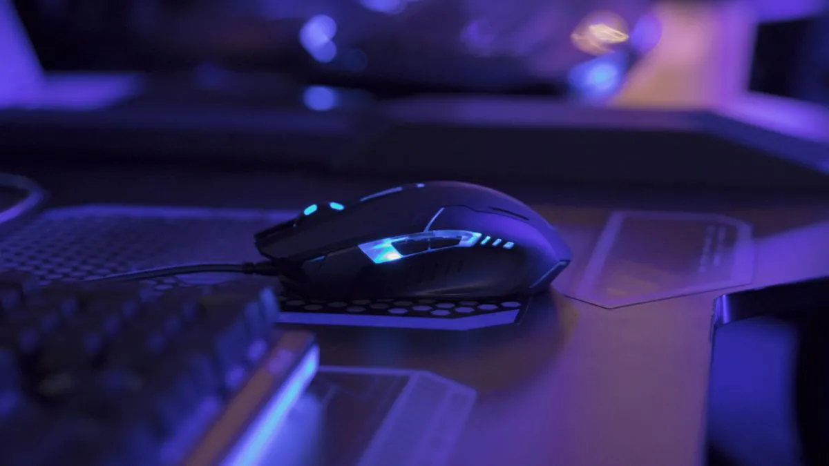 Chuột gaming với đèn LED phát sáng trên bàn làm việc trong môi trường ánh sáng xanh, tượng trưng cho trải nghiệm chơi game.